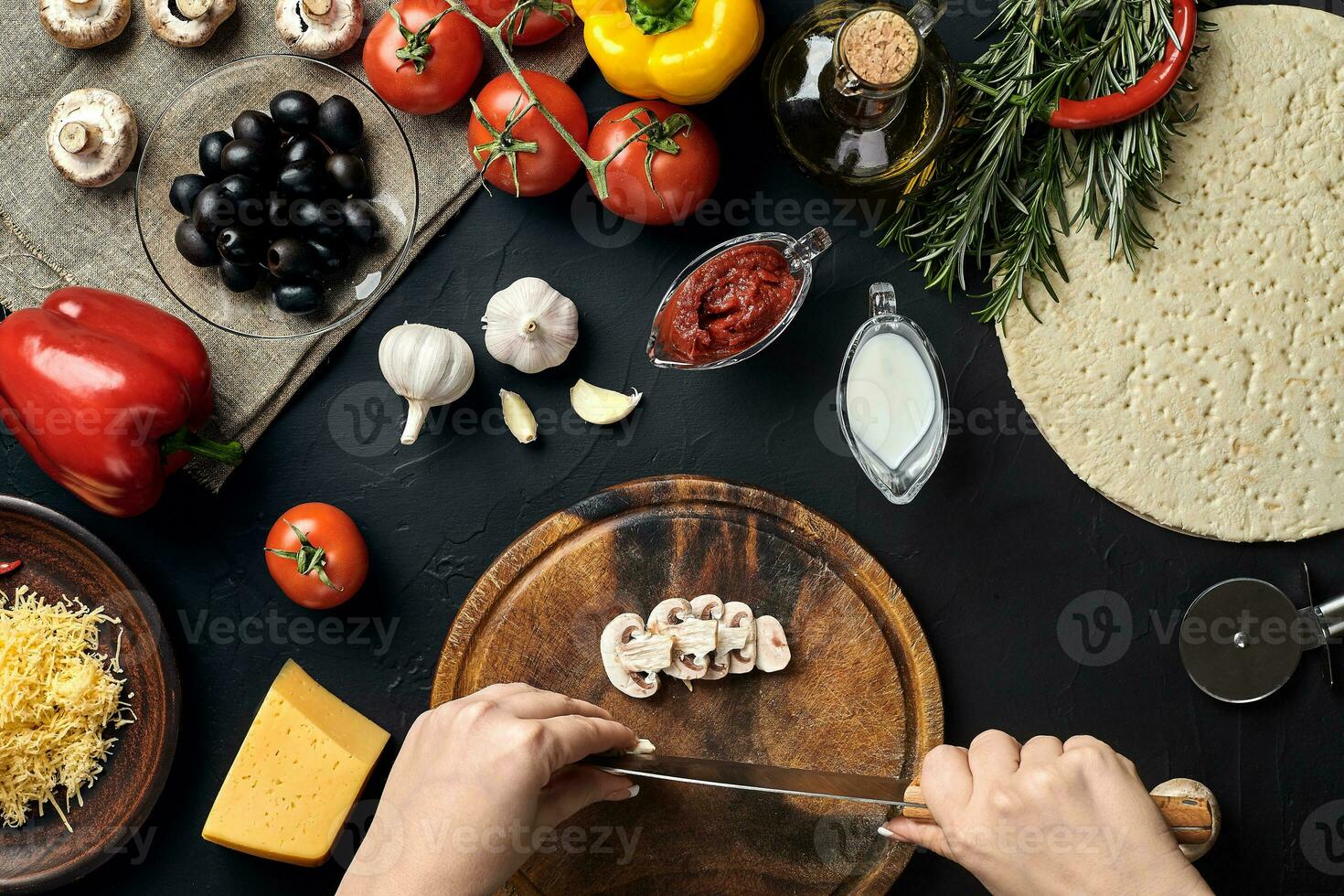Female hand cut mushrooms on wooden board on kitchen table, around lie ingredients for pizza vegetables, cheese and spices. photo