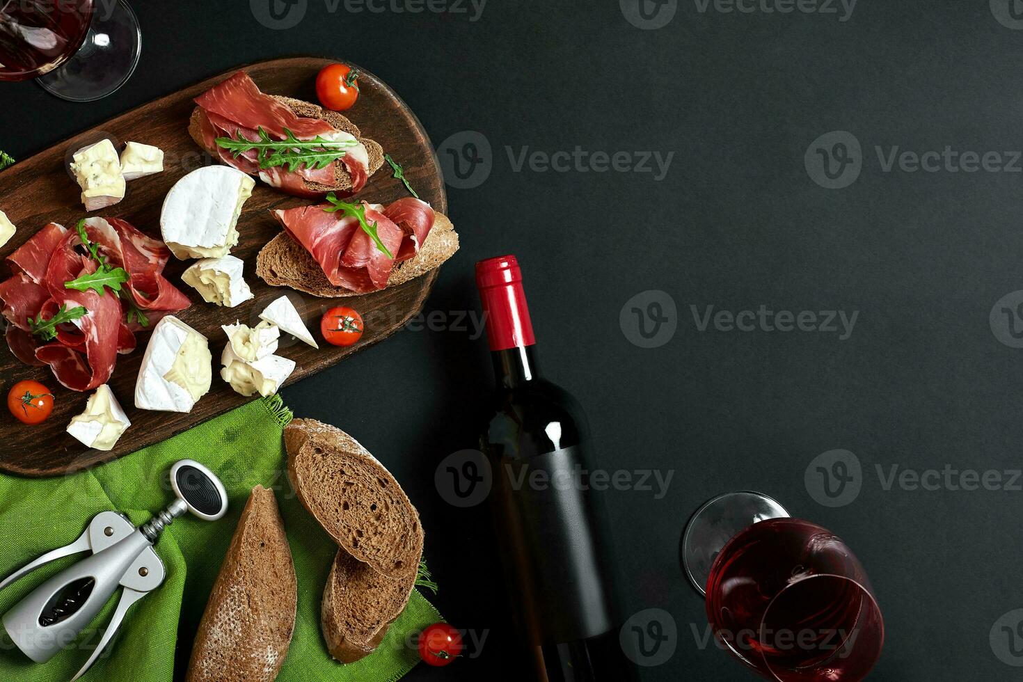 Delicious appetizer to wine - ham, cheese, baguette slices, tomatoes, served on a wooden board, and glass with red wine on black surface photo