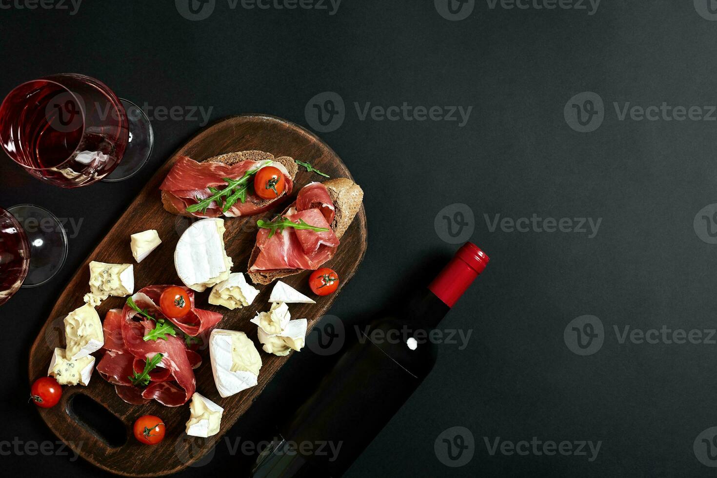 Delicious appetizer to wine - ham, cheese, baguette slices, tomatoes, served on a wooden board, and glass with red wine on black surface photo