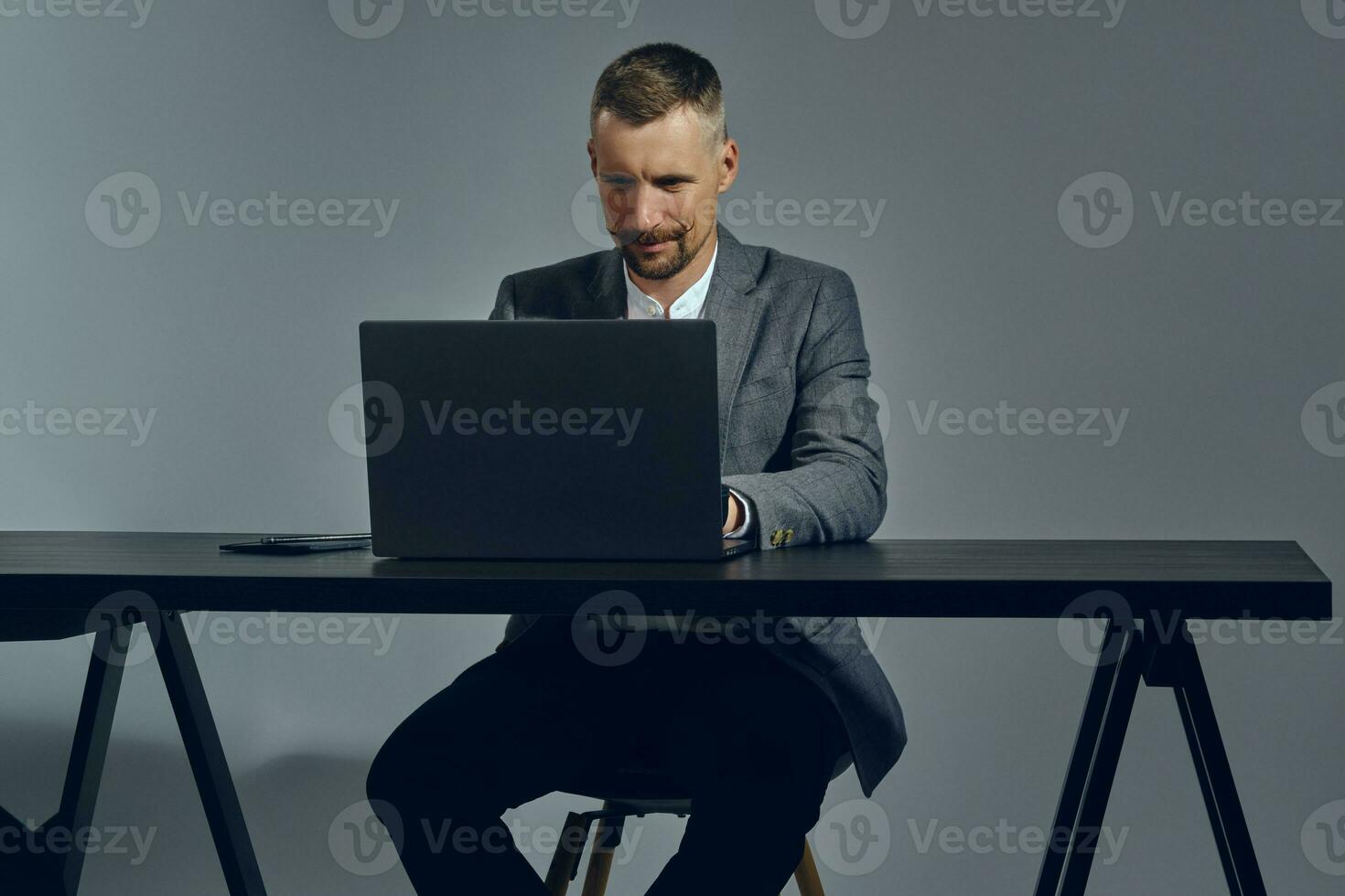 Bearded businessman with stylish mustache, dressed in a classic suit is working at laptop while sitting at table in office, gray background. photo