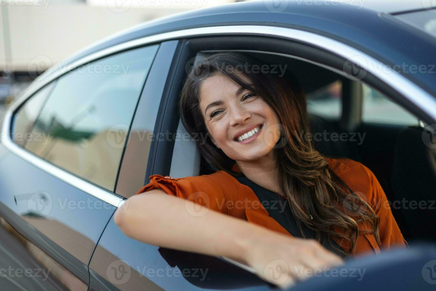 Young woman sitting in a car hand out of window. Happy woman driving a car and smiling. Portrait of happy female driver steering car with safety belt. Cute young lady happy driving car. photo