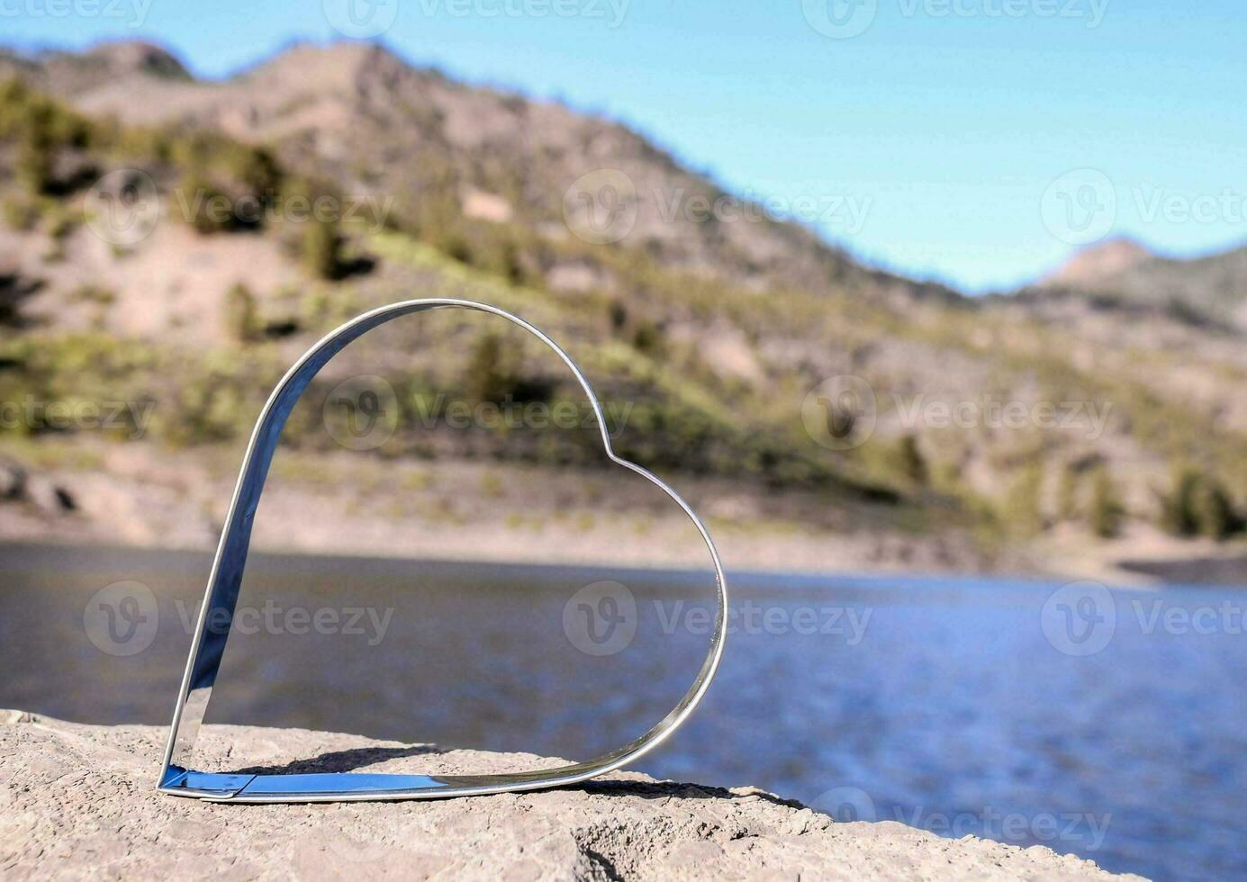 a heart shaped metal object sitting on a rock by a lake photo