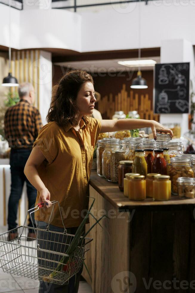 Woman holding shopping basket, buying ecofriendly bulk products in zero waste store designed to minimize plastic usage. Green living customer looking for nutritious groceries photo