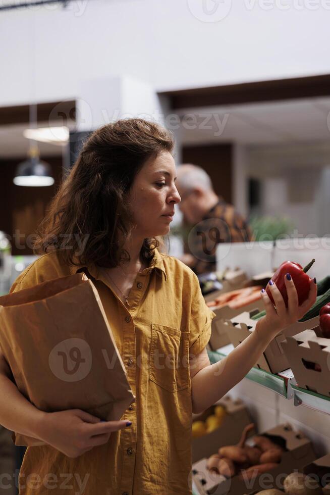 Woman in zero waste shop purchasing farm grown vegetables, picking ripe bell peppers. Client in plastic free local grocery shop looking to buy nutritious food, using decomposable paper bag photo