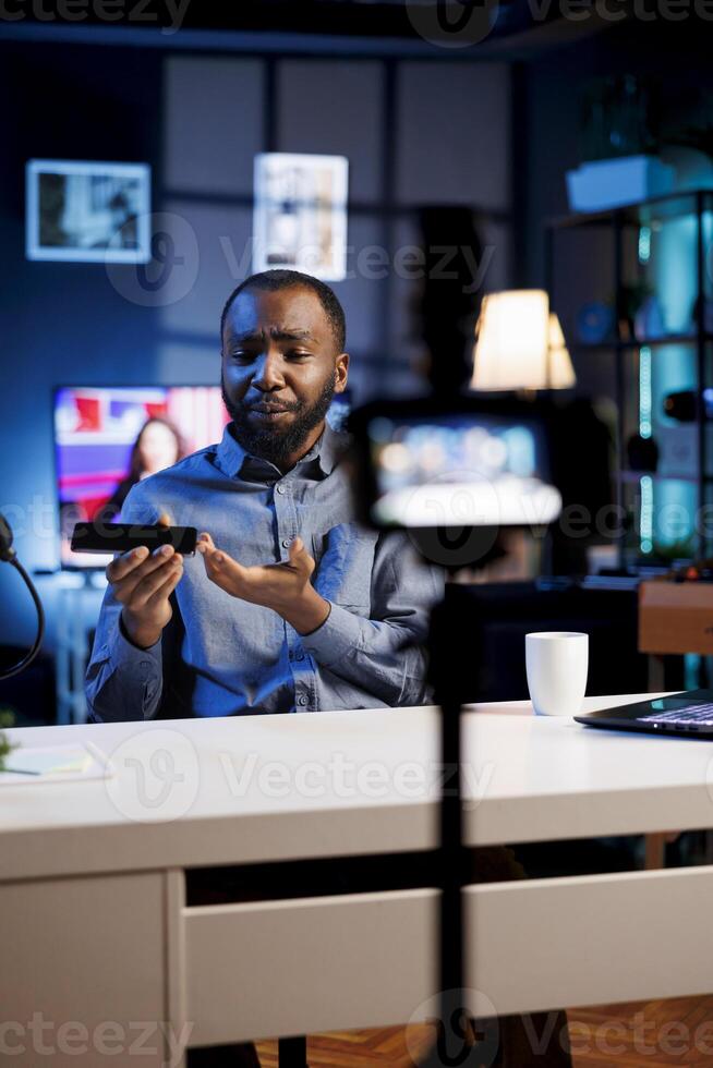 African american content creator analyzing newly launched smartphone received from sponsor, doing honest review for online streaming channel. Internet show host reviewing mobile phone brand photo