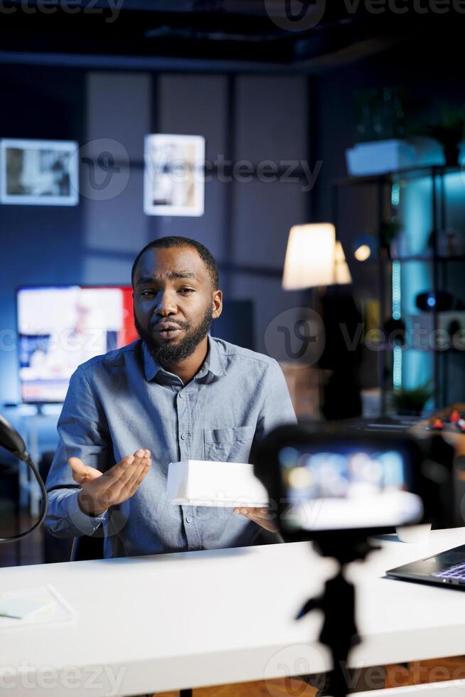 African american internet show host being sponsored by partnering brand to do unboxing video on streaming platform, gifting one to viewers. Man does influencer marketing, offering product as giveaway photo