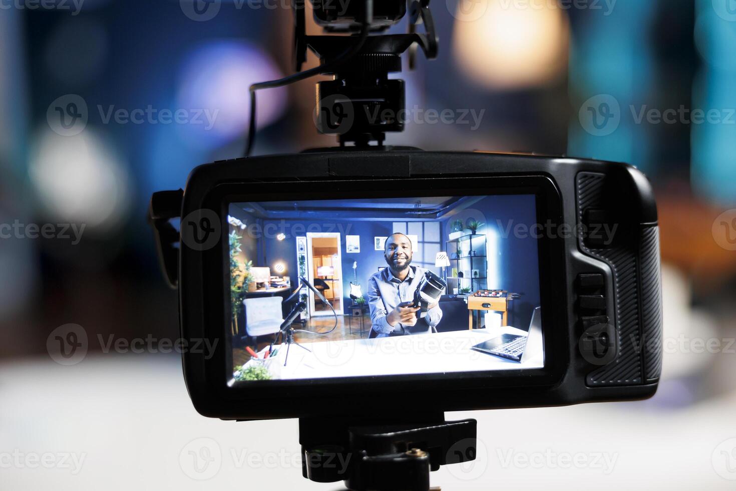 Focus on professional video production camera used for capturing footage of tech engineer in blurry background filming virtual reality product review for online audience, showcasing VR headset photo