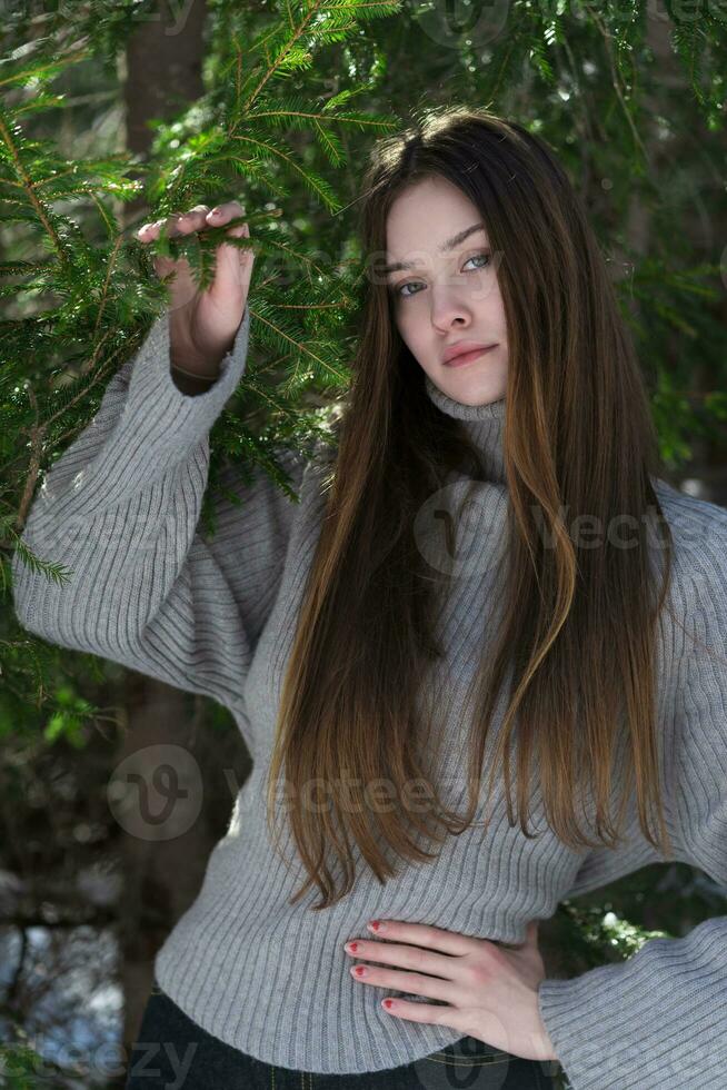 Teenager girl looking at camera, holding hand on waist, other raised and holds branch in pine forest photo