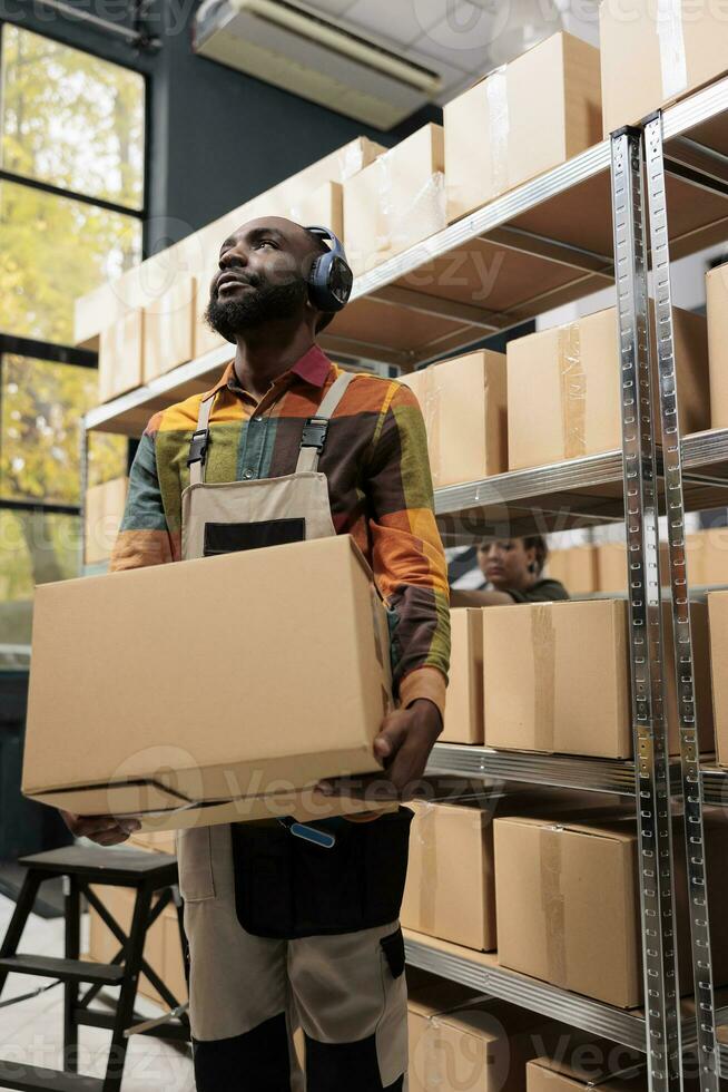 Supervisor working at merchandise distribution in storage room, carrying cardboard boxes while listening music. Storehouse manager wearing headphones, preparing customers packages for delivery photo