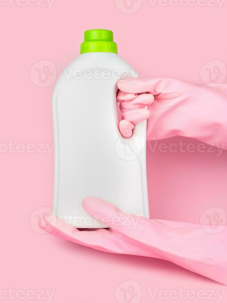 A hands in a pink rubber gloves holds a bottle of laundry conditioner on a pink background. Mockup. Close-up. Selective focus. photo