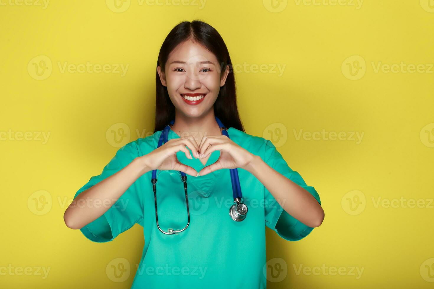 un asiático mujer vistiendo un del doctor uniforme poses con su manos en el forma de un corazón en pie en frente de un amarillo antecedentes. foto