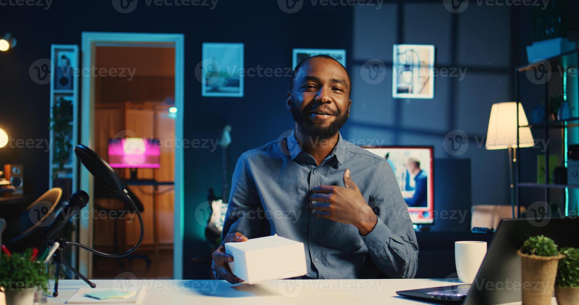 Tech content creator filming packaged product unboxing in neon lights personal home studio. BIPOC cheerful influencer showing sponsoring brand merchandise, urging subscribers to purchase photo