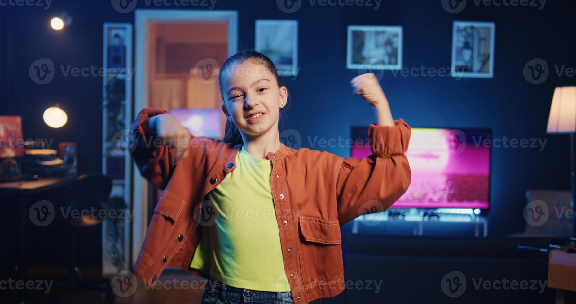 Happy child in apartment illuminated by RBG lights filming dance tutorial for other children watching her online social media content. Kid influencer in studio recording video targeting gen Z fans photo