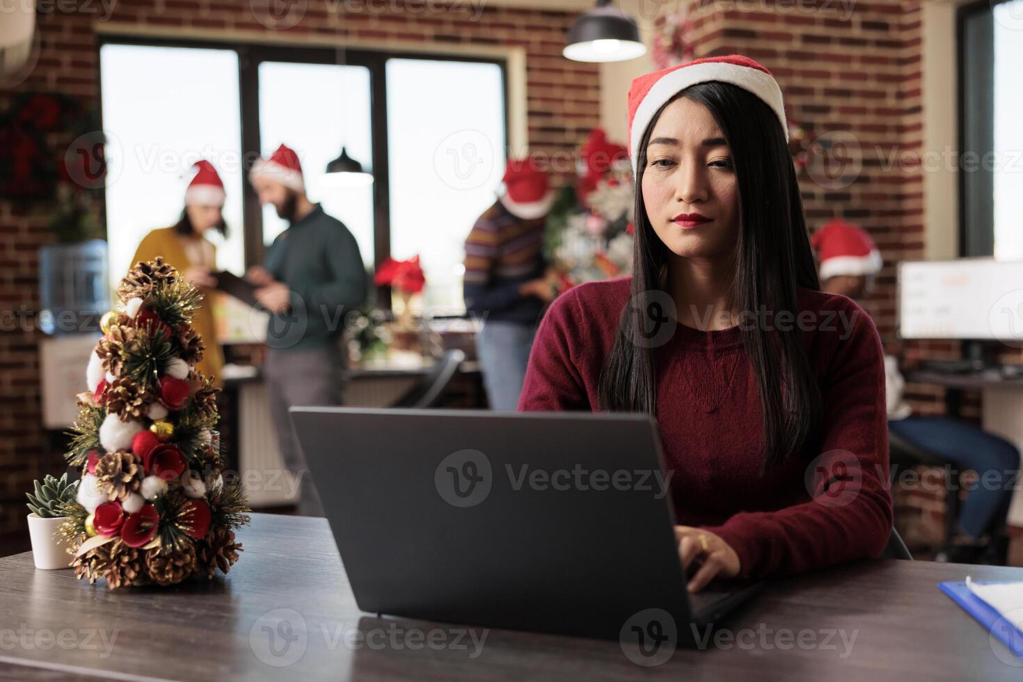 Asian woman corporate worker in santa hat working on project at winter seasonal holiday in decorated office. Company employee using typing on computer at festive coworking space photo