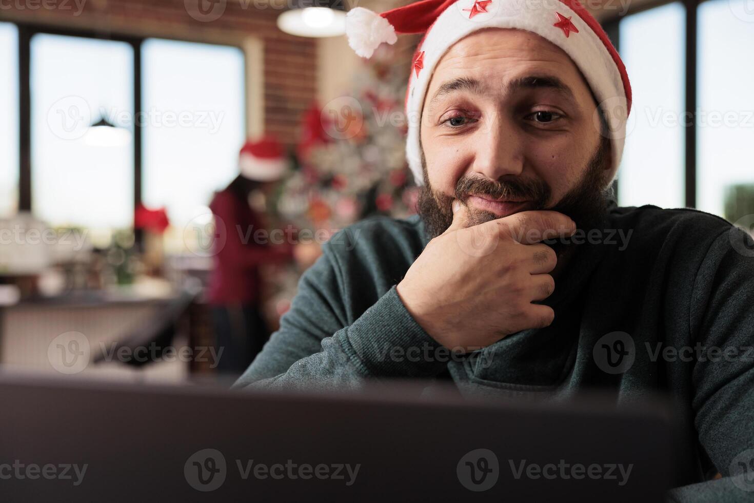Smiling worker in santa hat rubbing chin while reading research report on laptop in office. Caucasian man company employee working on computer at christmas eve during winter holiday photo