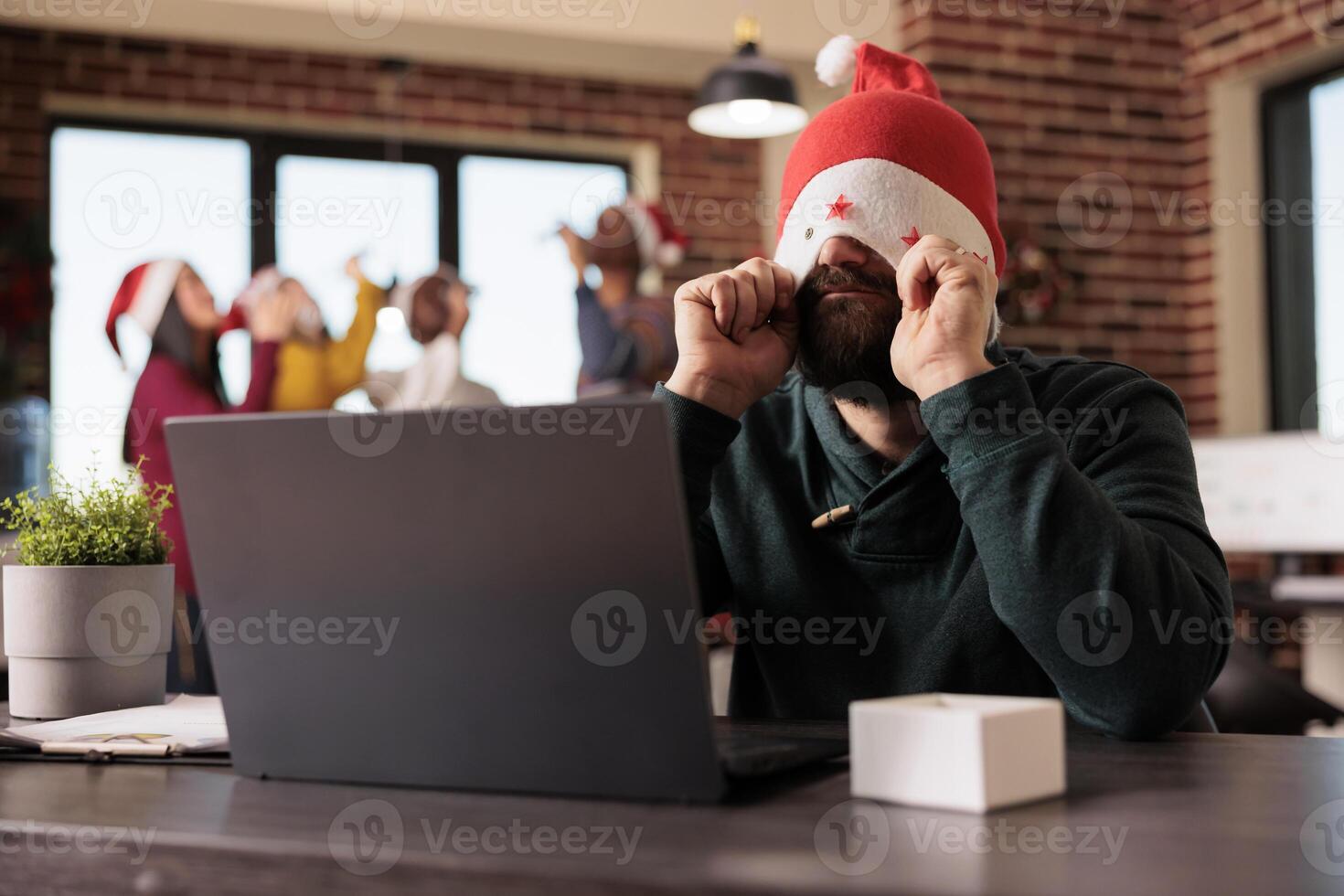 Frustrated tired employee putting santa hat on eyes while working overtime during christmas season in office. Startup company worker managing tasks workload during winter holiday photo