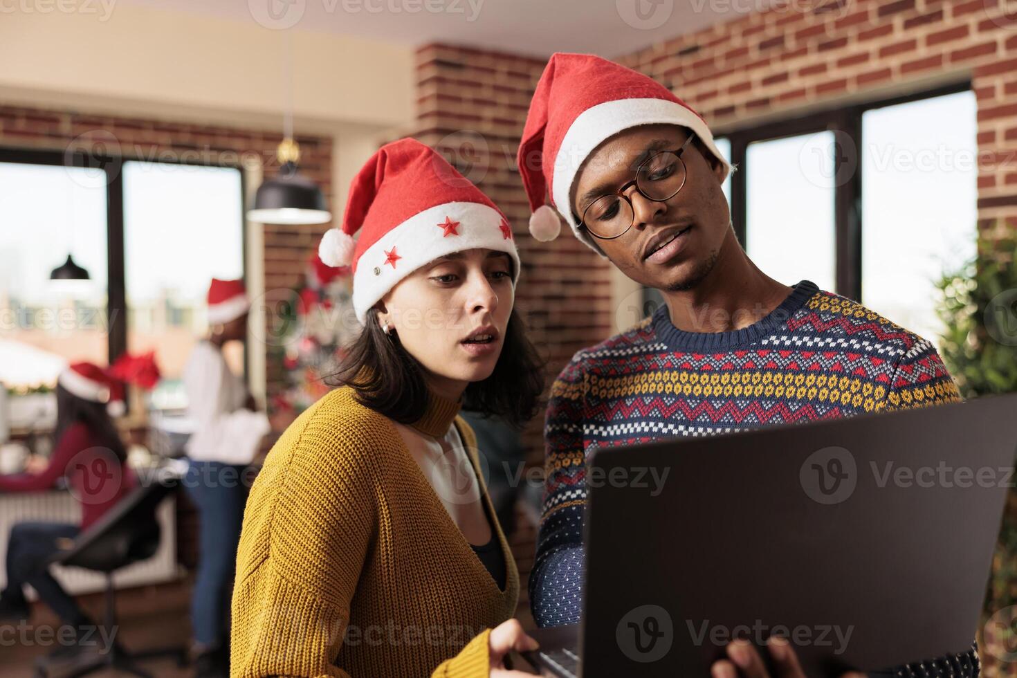 Coworkers in santa hats holding laptop and checking business research report in office during christmas holiday. Diverse colleagues managing workload at xmas eve in workplace photo