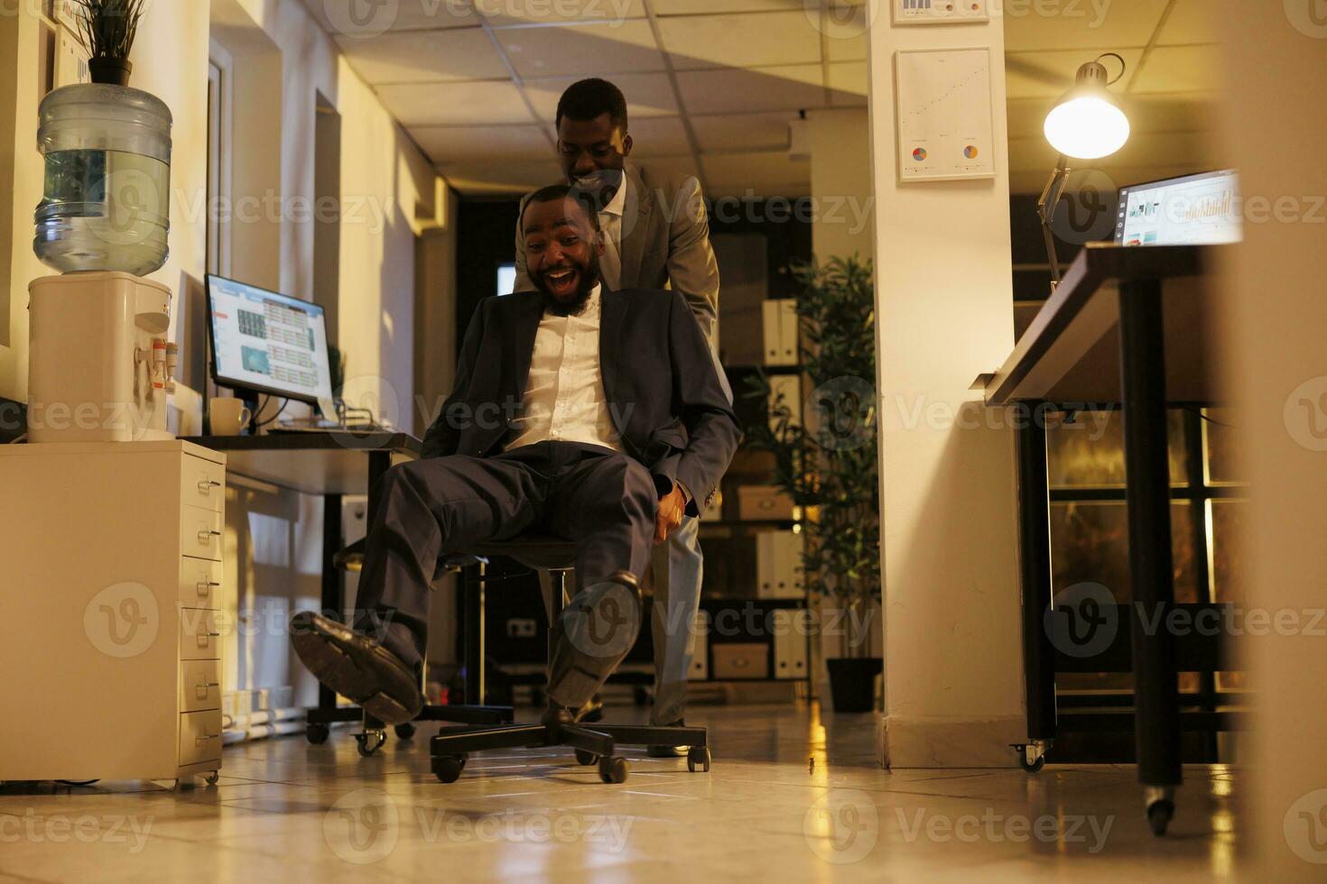 Cheerful smiling businessmen having sports race competition in modern startup office, taking break from work and having fun. Playful businessmen in formal suit enjoying riding desk chair photo