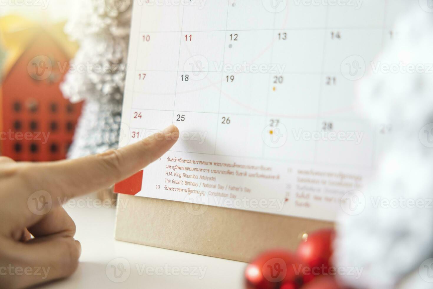 A woman's fingers pointing at the calendar on December 25th, Christmas Day by the festive scene of beautiful Christmas tree adorned, with Christmas balls, pine cones. concept about Important day. photo
