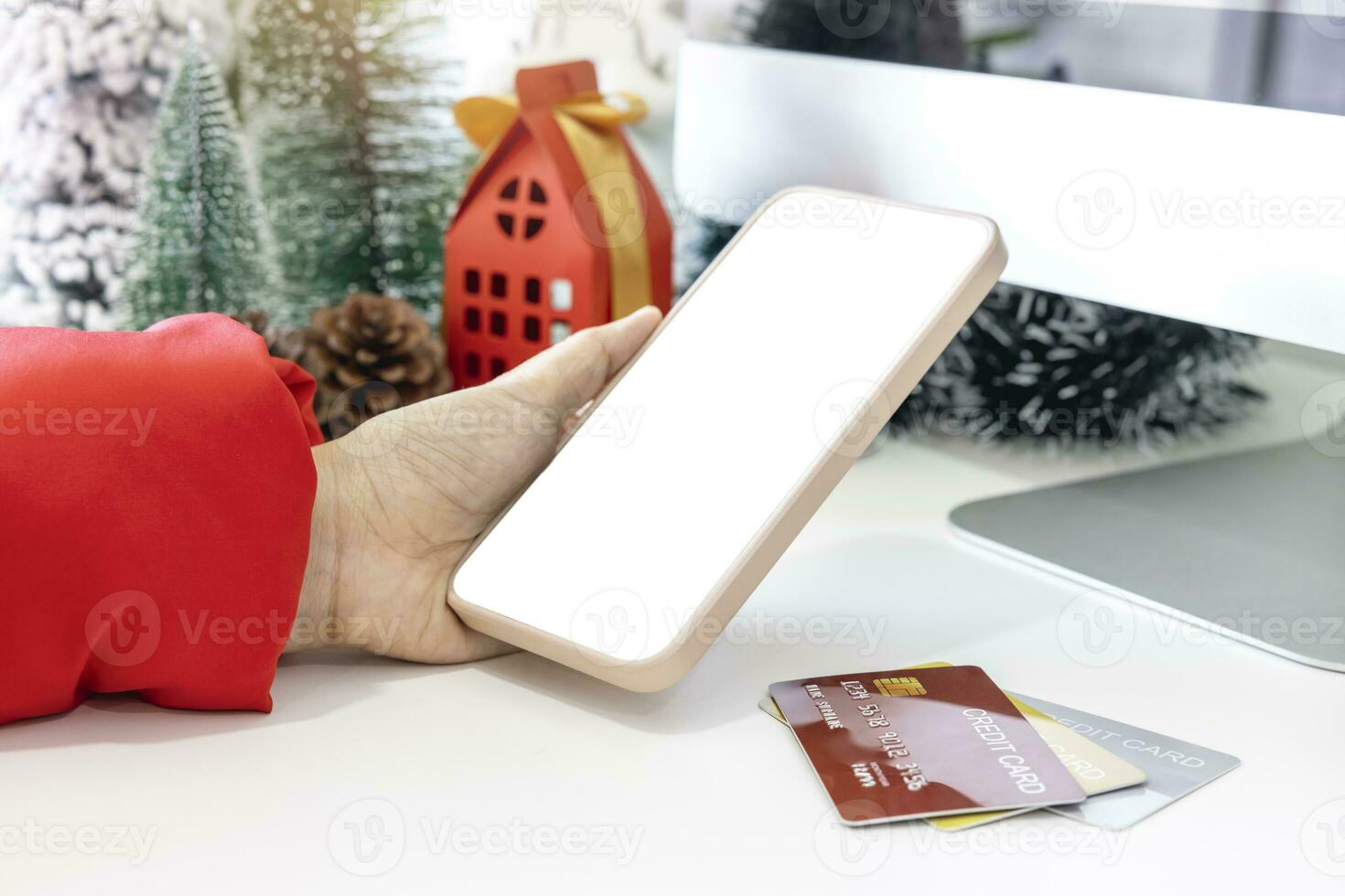 Gentle hands cradle a smartphone, empty screen by festive scene a beautiful Christmas tree adorned with credit cards, along with Christmas balls, pine cones, and red house gift box tied golden ribbon. photo