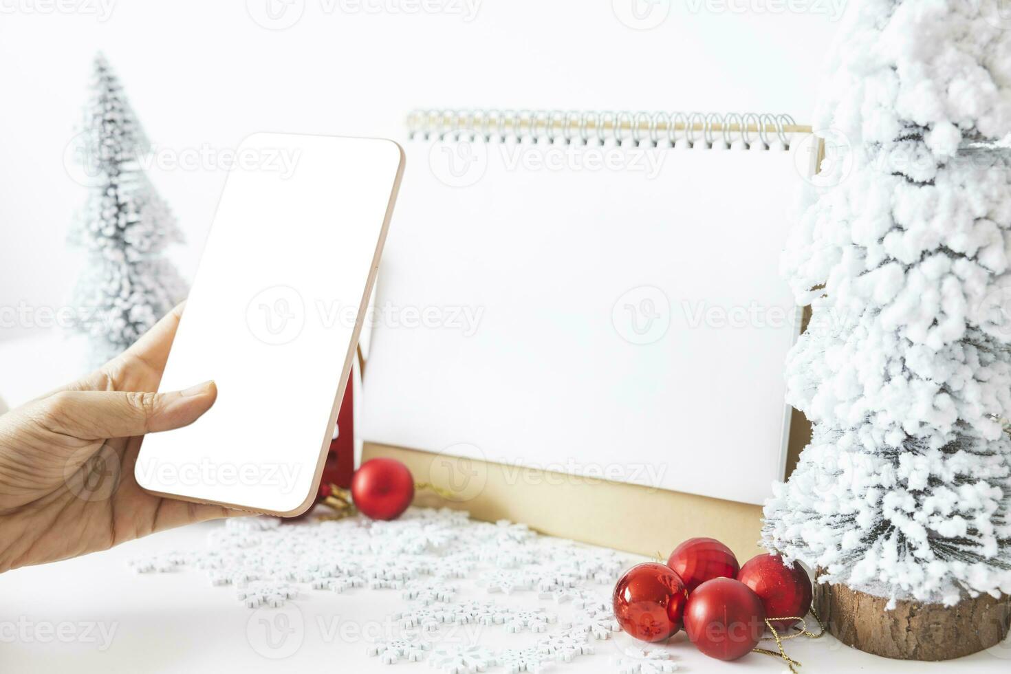 Gentle hands cradle a smartphone with a white screen mock-up and White calendar with a festive scene of a beautiful Christmas tree adorned, with Christmas balls, pine cones, and a red house gift box. photo