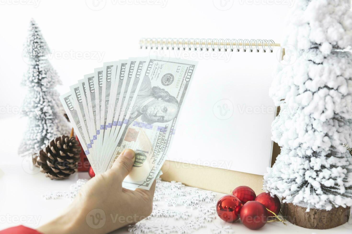 Gentle hands cradle money with a white screen mock-up and White calendar with a festive scene of a beautiful Christmas tree adorned, with Christmas balls, pine cones, and a red house gift box. photo