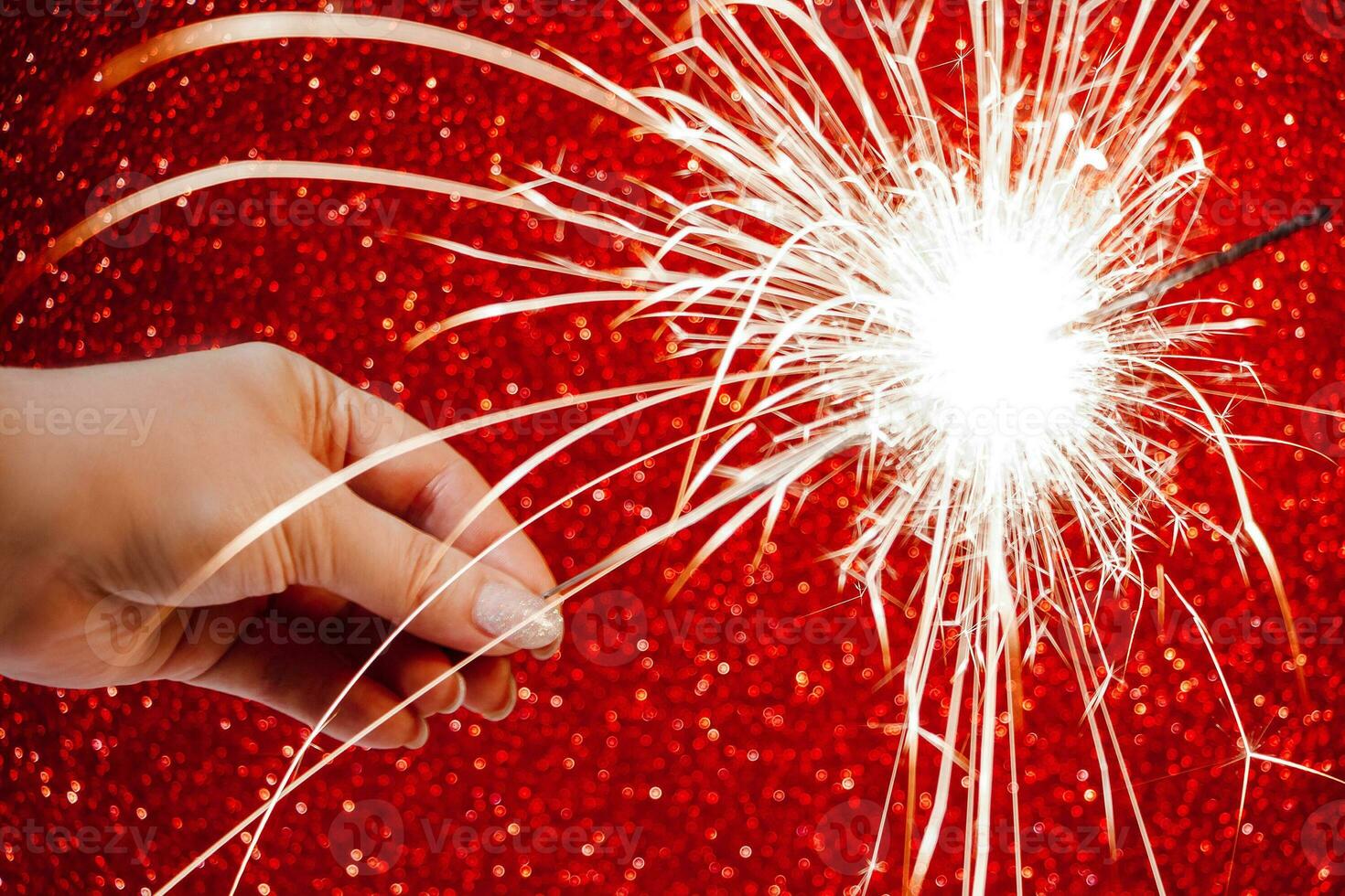 New Year celebration. Womans hand holds bright sparkler on red background. Christmas night. Bright sparks of fire and bokeh. photo