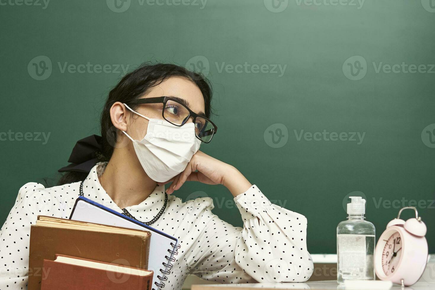 a young teacher wearing a face mask sits at a desk with books photo