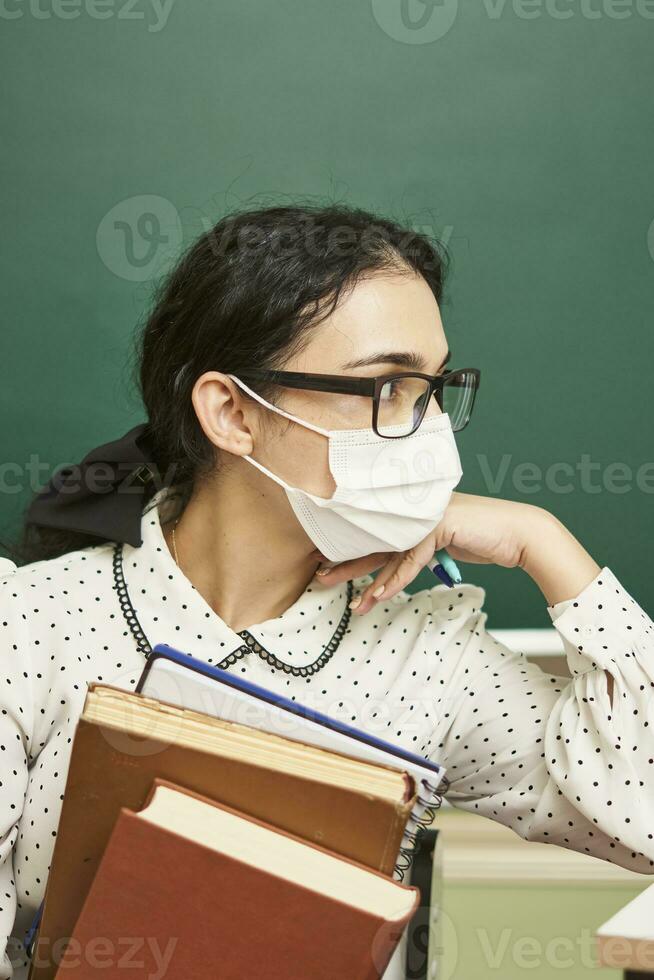 a young teacher wearing a face mask sits at a desk with books photo