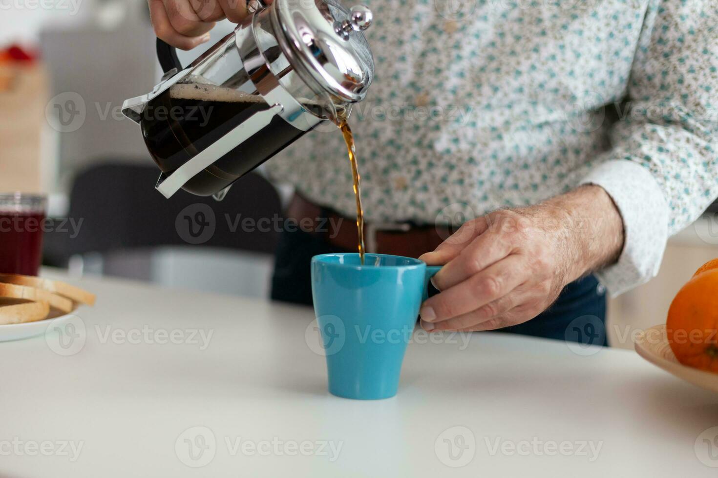 Senior man using french press and pouring hot drink after preparing coffee in kitchen for breakfast. Elderly person in the morning enjoying fresh brown cafe espresso cup caffeine from vintage mug photo