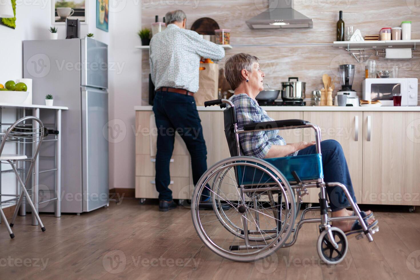 Paralysed senior woman in wheelchair feeling unhappy sitting in the middle of kitchen while husband unpacking grocery bag. Old handicapped lady after injury and rehab, depressed invalid full of sorrow photo