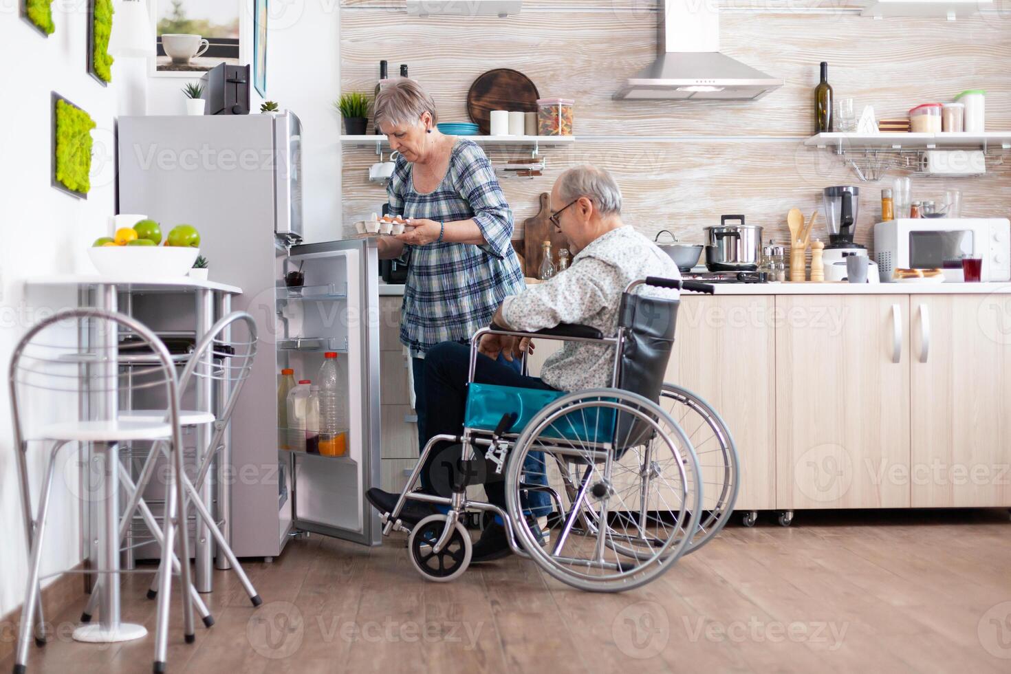 Senior woman preparing breakfast for handicapped husband taking eggs carton from refrigerator , living with man with walking disabilities. Disabled senior male in wheelchairhelping his wife in kitchen. photo
