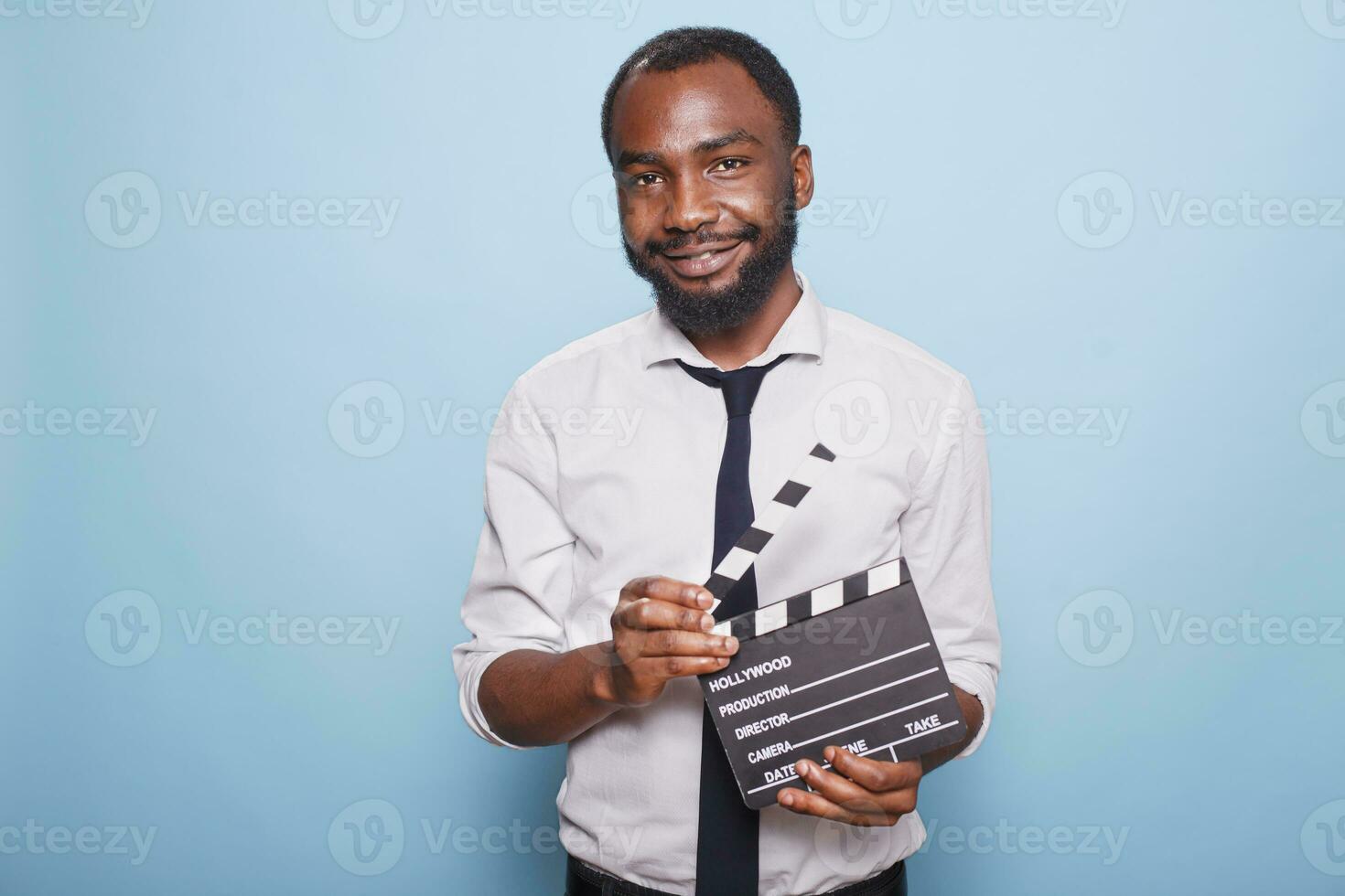 African American movie director holding open clapperboard in hands looking confident while posing for camera. Young successful filmmaker grasping clapper standing proud against blue background. photo