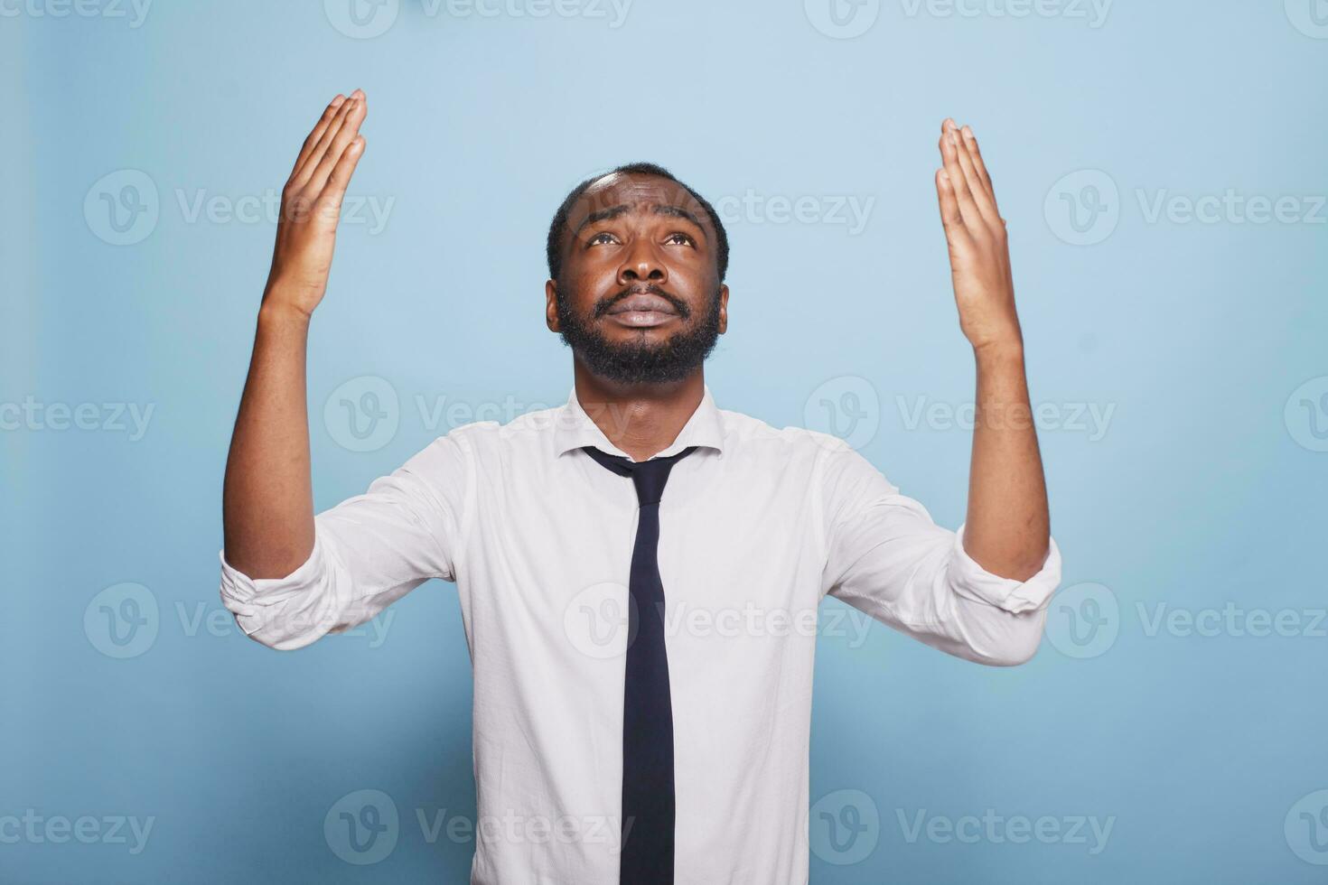 preocupado negro empresario es Orando y levantamiento su manos en un humilde gesto hacia el cielo. africano americano empleado vistiendo un negro Corbata y un blanco camisa suplica para perdón foto
