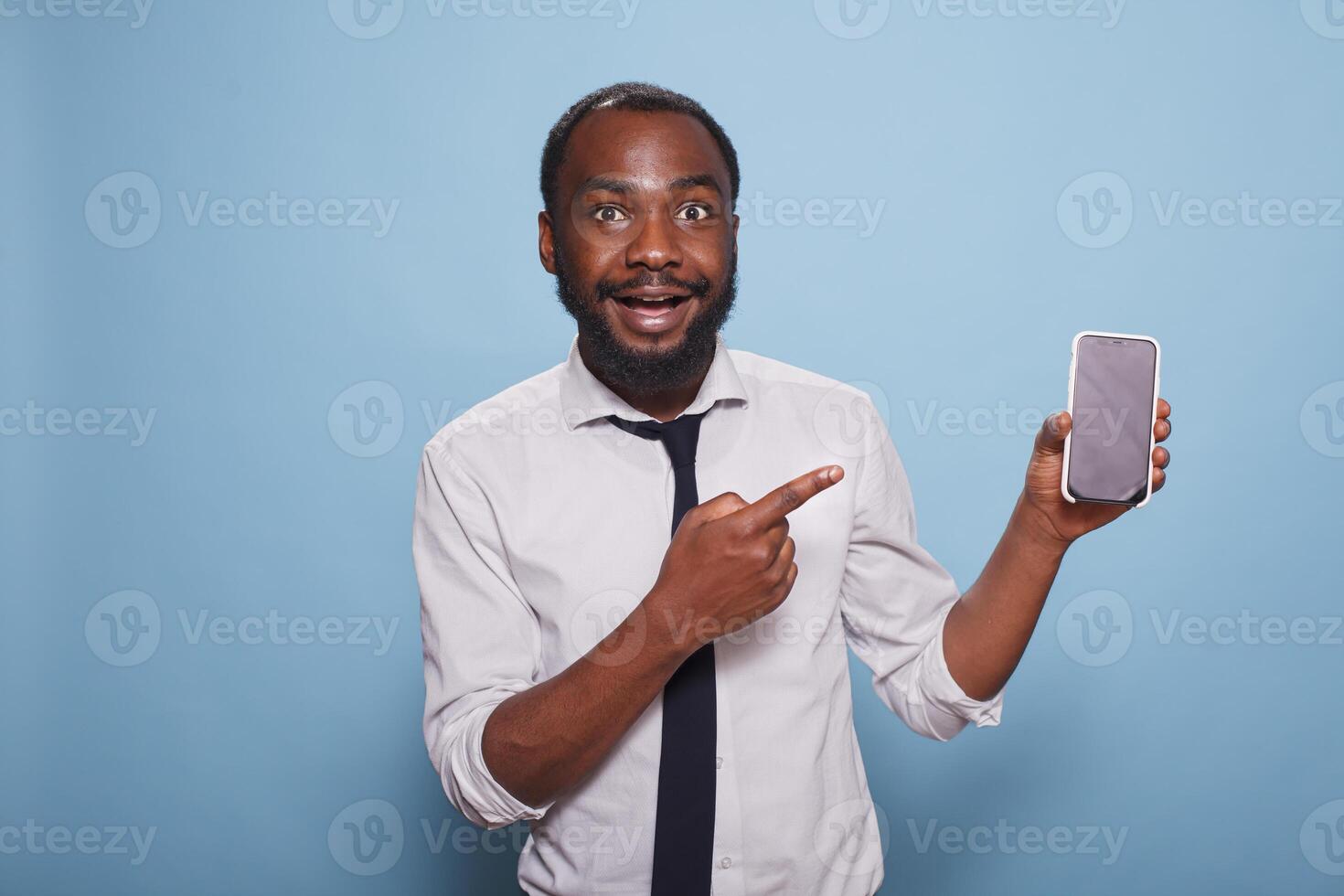 Enthusiastic black man pointing at smartphone blank screen showing mobile feature of mobile game or app. Excited individual using index finger to enhance features of digital device. photo