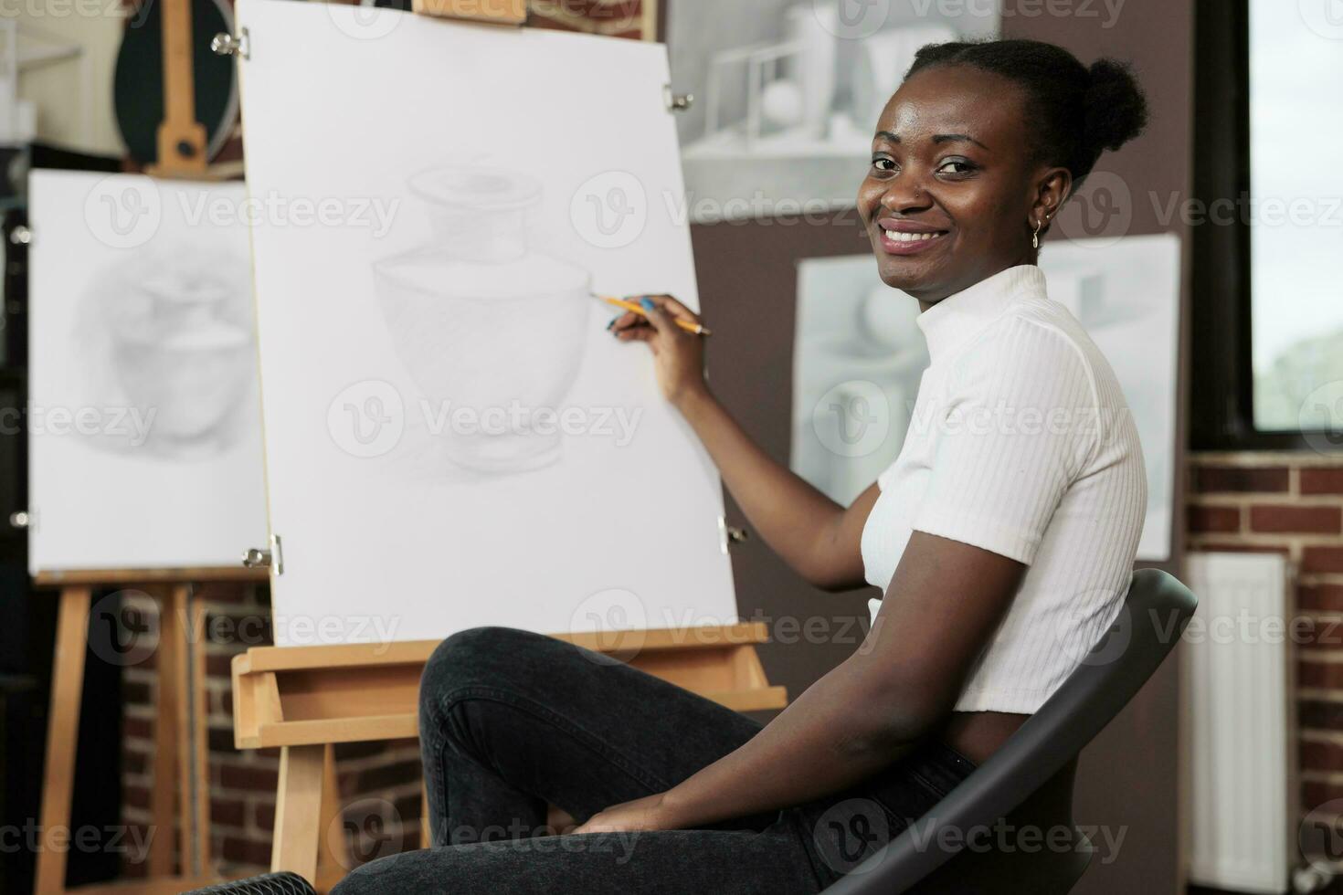 Happy woman sitting on chair at easel and smiling at camera, holding pencil creating masterpiece during group drawing class. Creative leisure activities and mental health concept photo