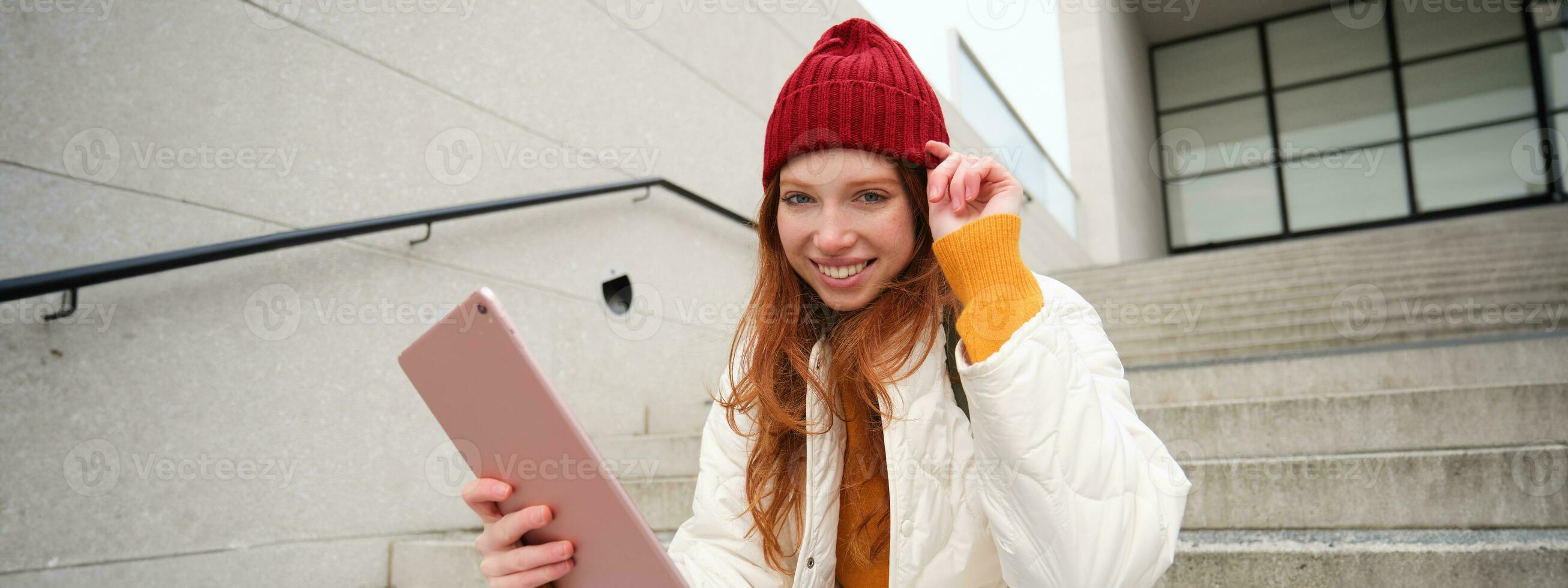 Young stylish girl, redhead female students sits on stairs outdoors with digital tablet, reads, uses social media app on gadget, plays games while waits on street photo