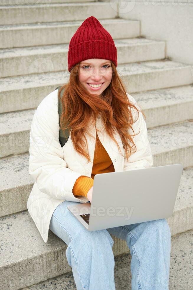 Smiling redhead girl, student sits on stairs outdoors and uses laptop, connects to public wifi in city and works on project, uses internet on computer photo