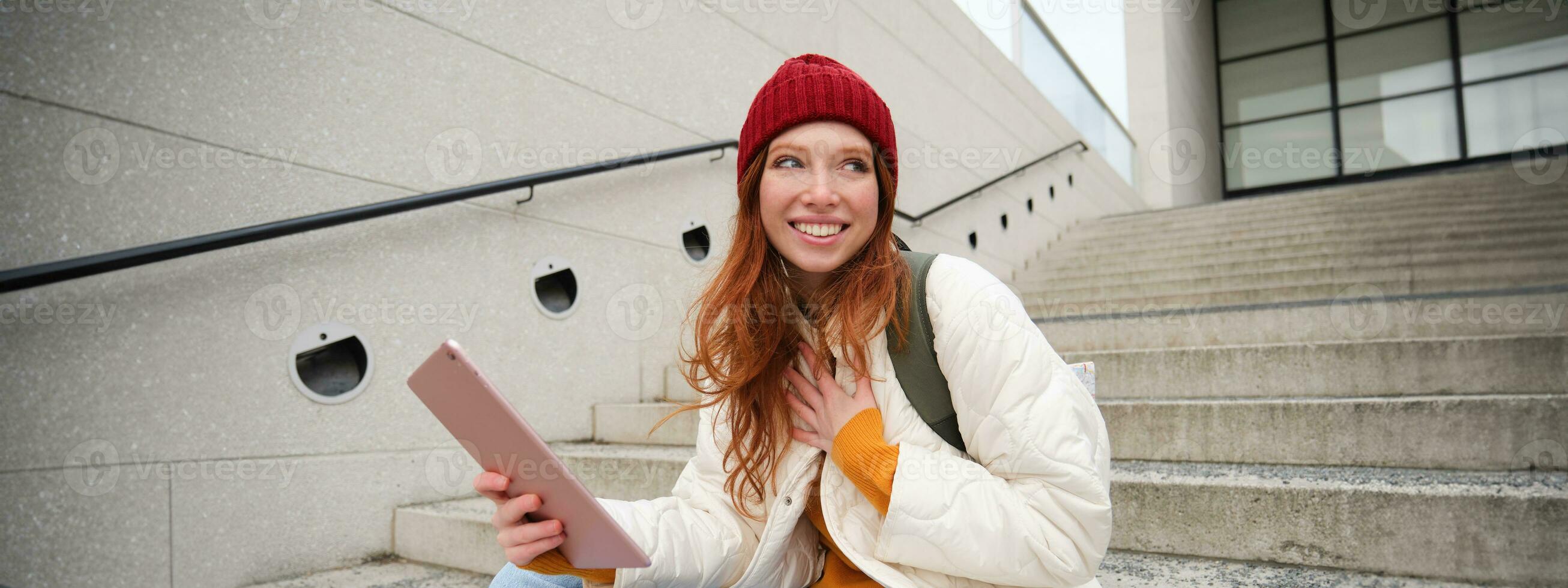 Young stylish girl, redhead female students sits on stairs outdoors with digital tablet, reads, uses social media app on gadget, plays games while waits on street photo