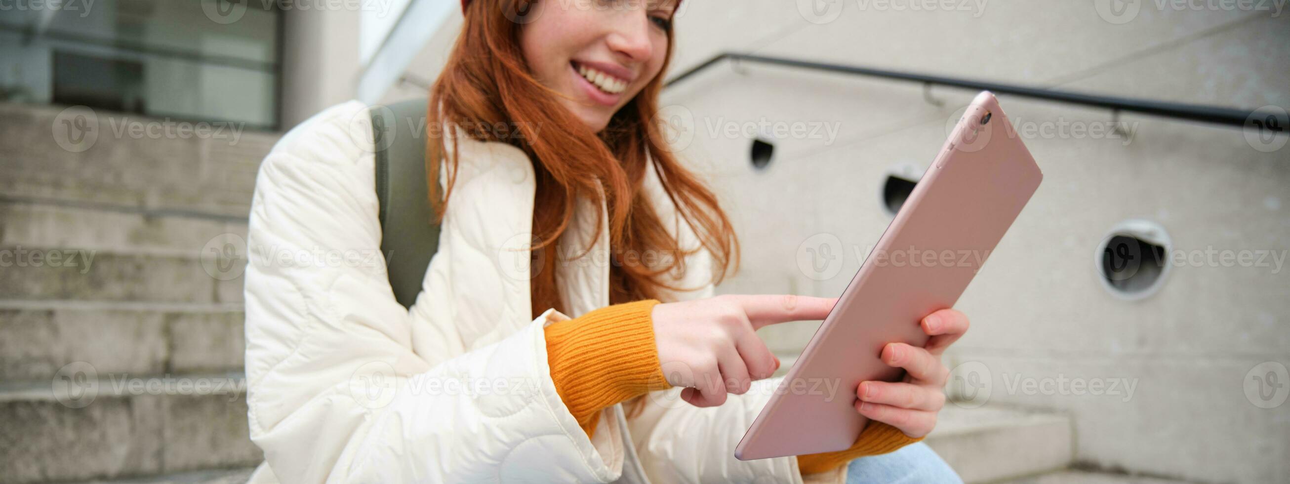 Happy stylish redhead girl, student in red hat, holds digital tablet, uses social media app, searches something online, connects to wifi photo