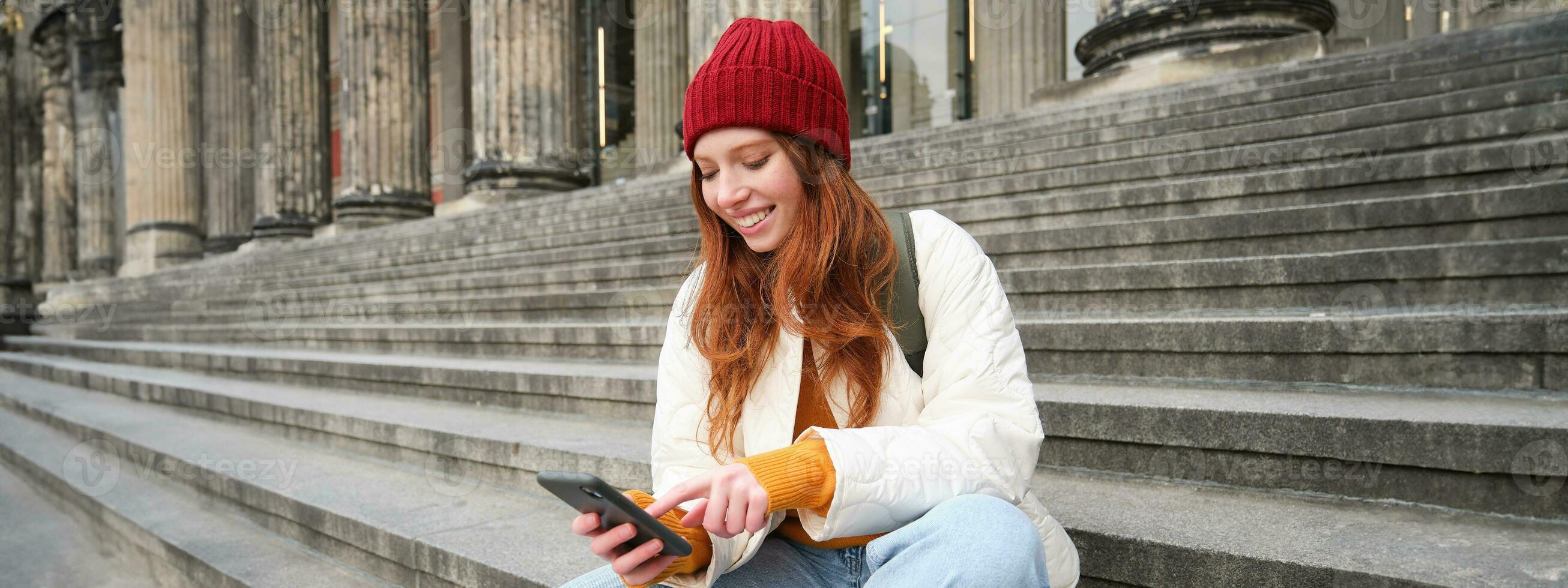 Portrait of young urban girl in red hat, sits on stairs near museum, holds mobile phone, connects to public wifi and surfs net, uses smartphone apps photo