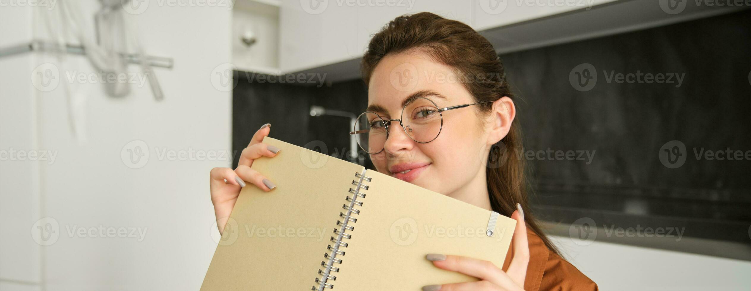 Close up portrait of smiling beautiful woman with notebook, making notes, writing in diary, studying at home, preparing homework at in kitchen photo