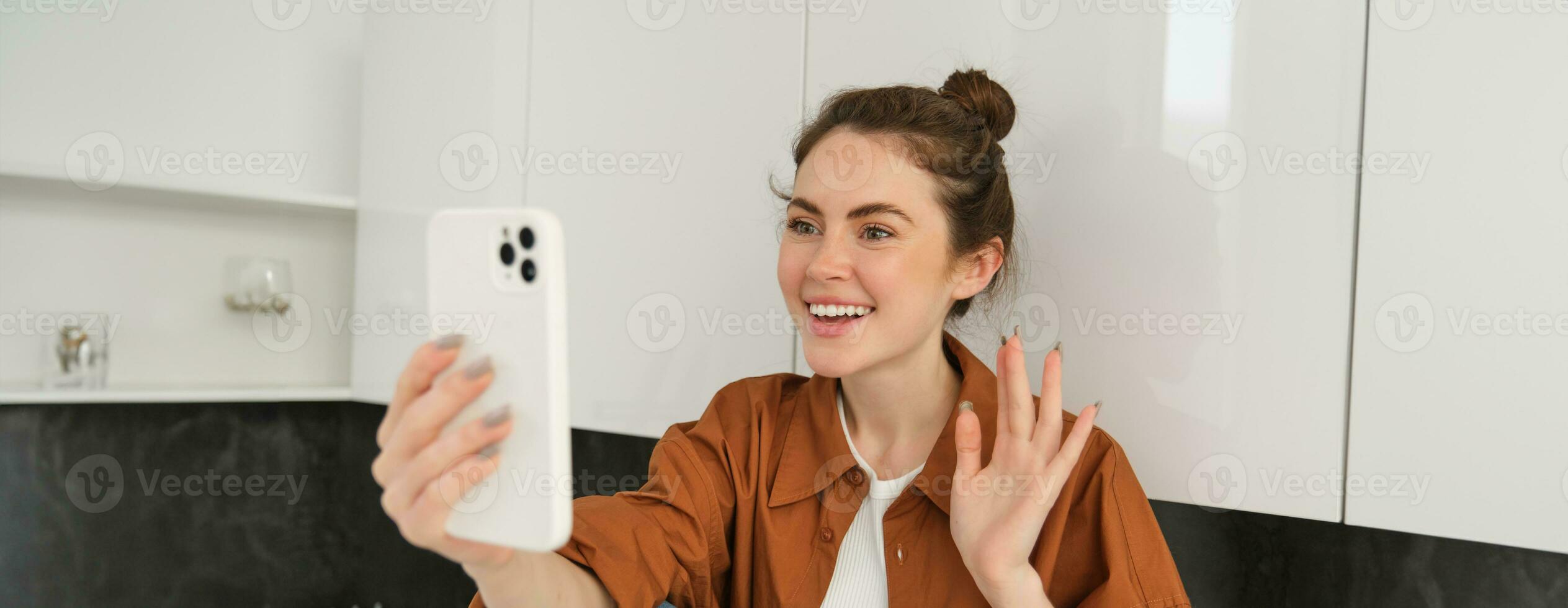 Portrait of young cheerful woman laughing and smiling during phone call, video chats with friend, sits on kitchen counter and talks to someone using smartphone app photo