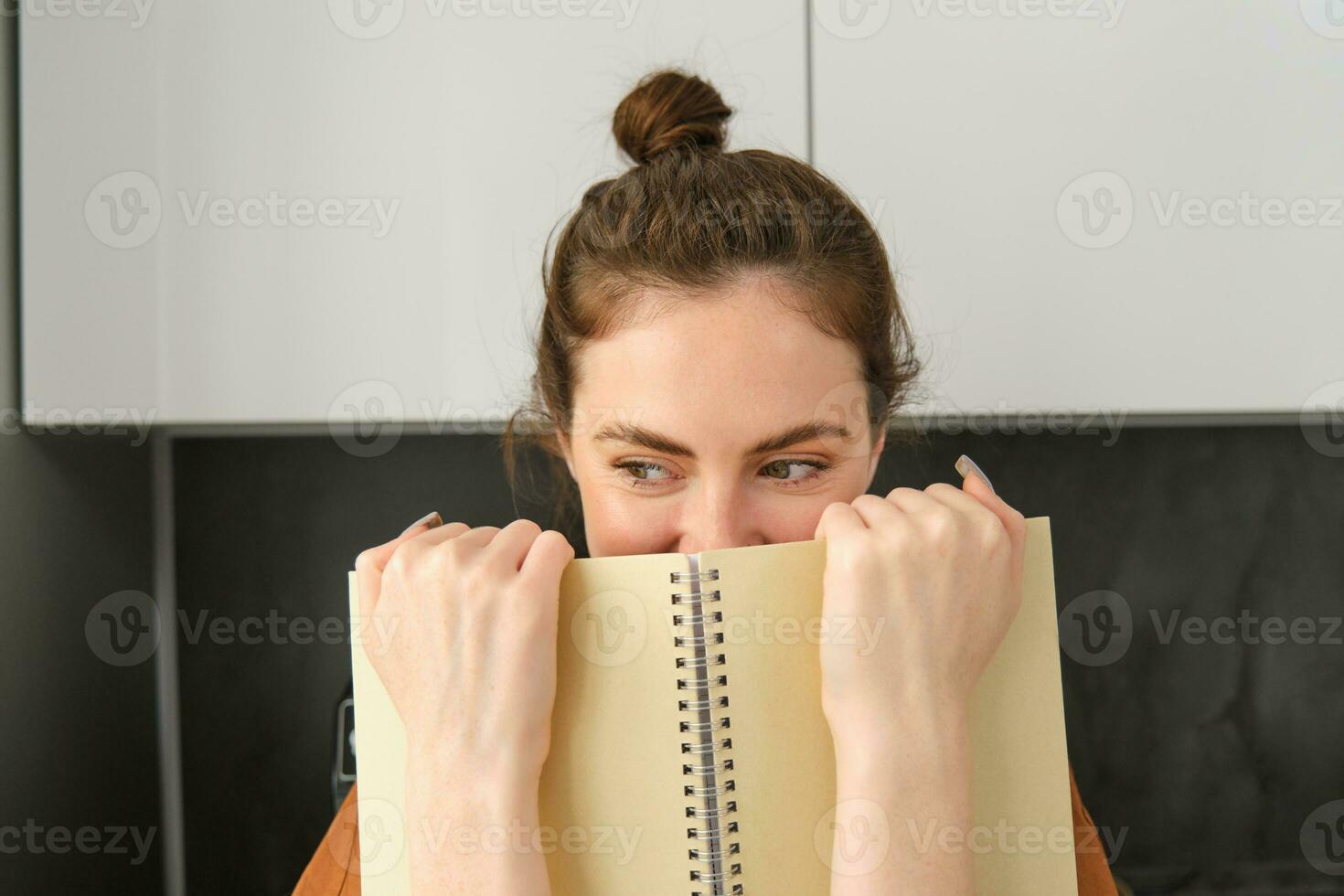 Cute woman hides face behind notebook, looking aside, smiling with eyes photo