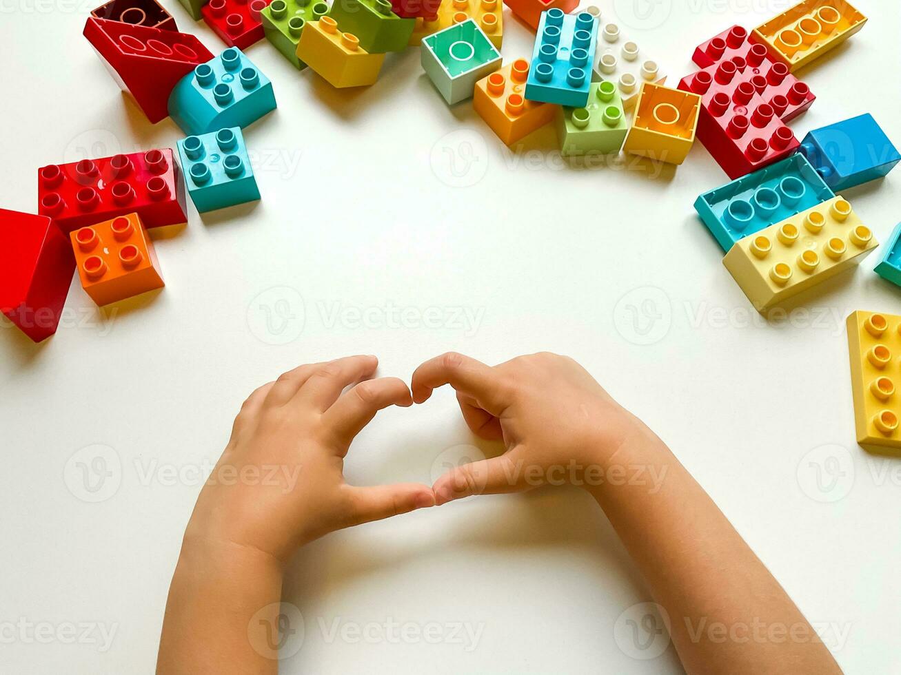 Child shows a heart against background of colorful building blocks on white background. Top view. photo