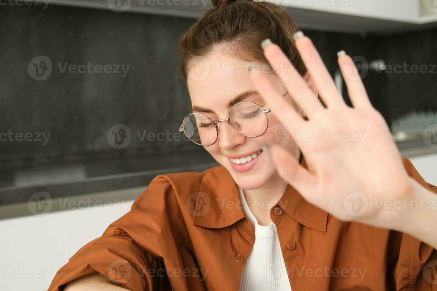 Close up portrait of cute young woman, covers, puts hand as block from camera and smiling, sitting in kitchen, laughing and smiling photo