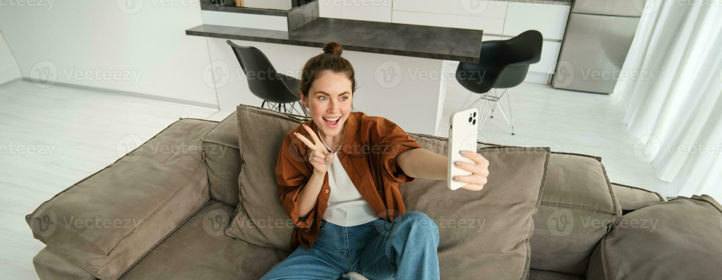 Portrait of cute smiling female model, takes selfie on smartphone app, posting photos on social media, using mobile phone camera, posing on sofa at home