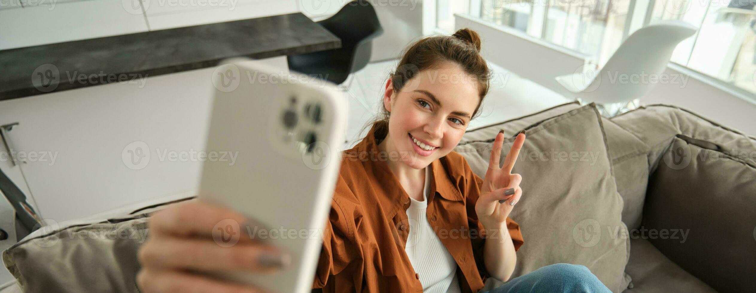 Positive and happy young woman taking selfie on smartphone, sitting on couch, showing peace, vsign gesture photo