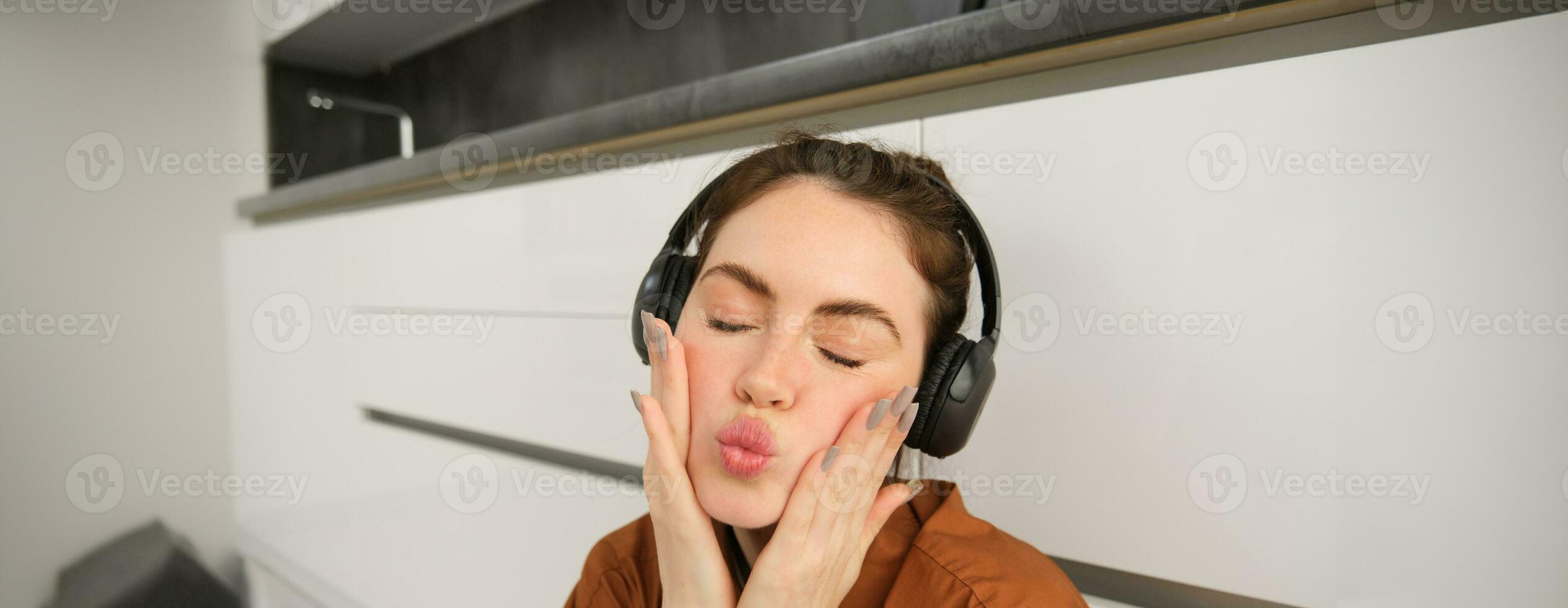 Beautiful young woman in wireless headphones, makes silly and cute faces, listens to music at home photo