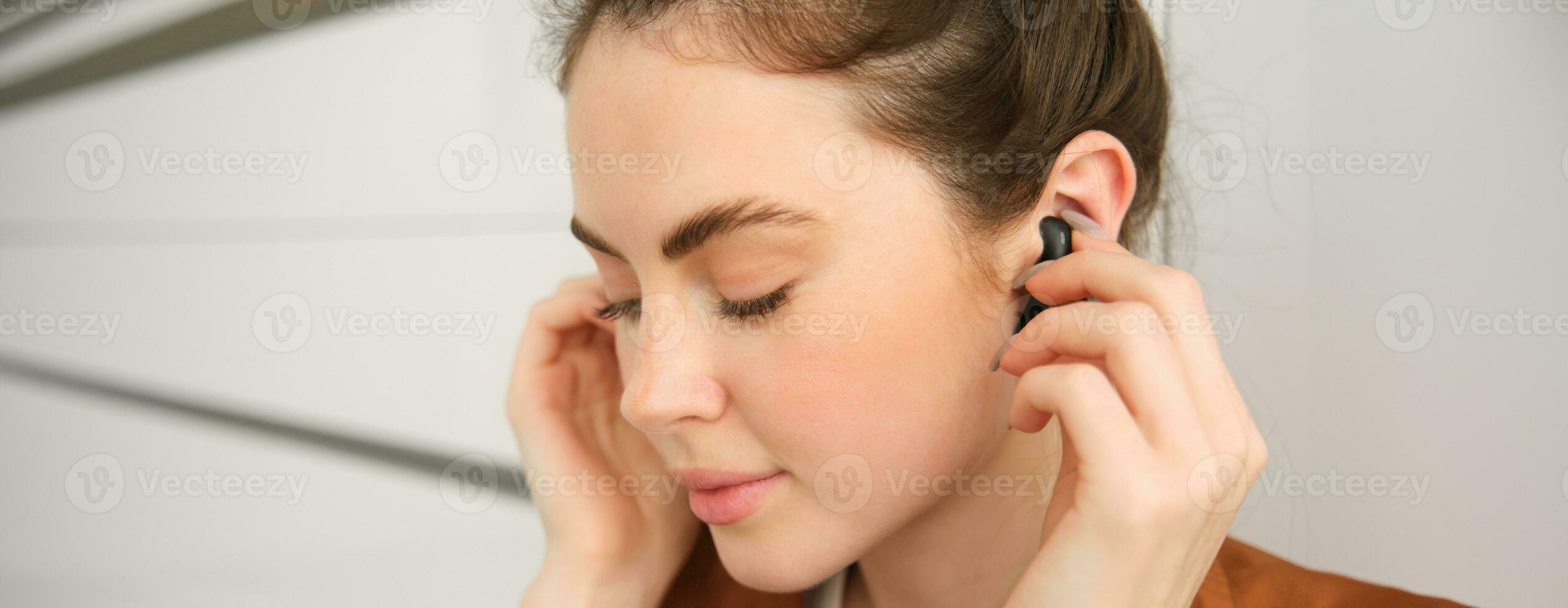 Close up portrait of tender, smiling and cute woman, puts on black wireless earphones in her ears, listens to music photo